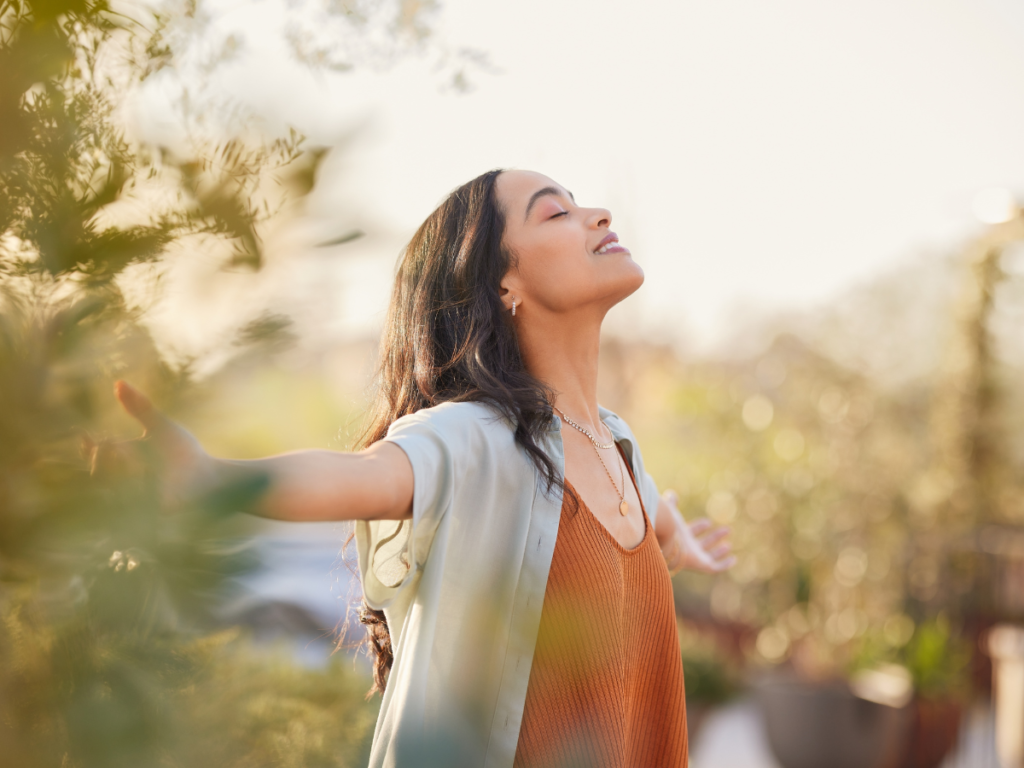 A woman standing outdoors with her arms open, eyes closed, and a serene expression, symbolizing relaxation and emotional regulation.