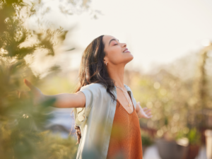 A woman standing outdoors with her arms open, eyes closed, and a serene expression, symbolizing relaxation and emotional regulation.